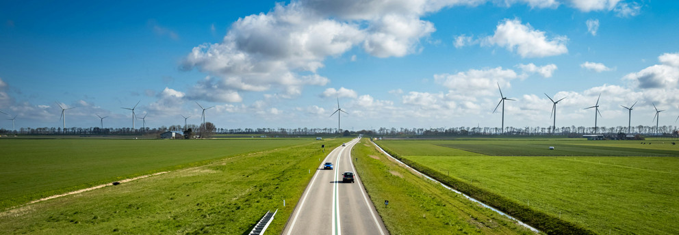 Panoramic image of a wind power plant on green farmland with cars driving on a crossing street on a sunny day